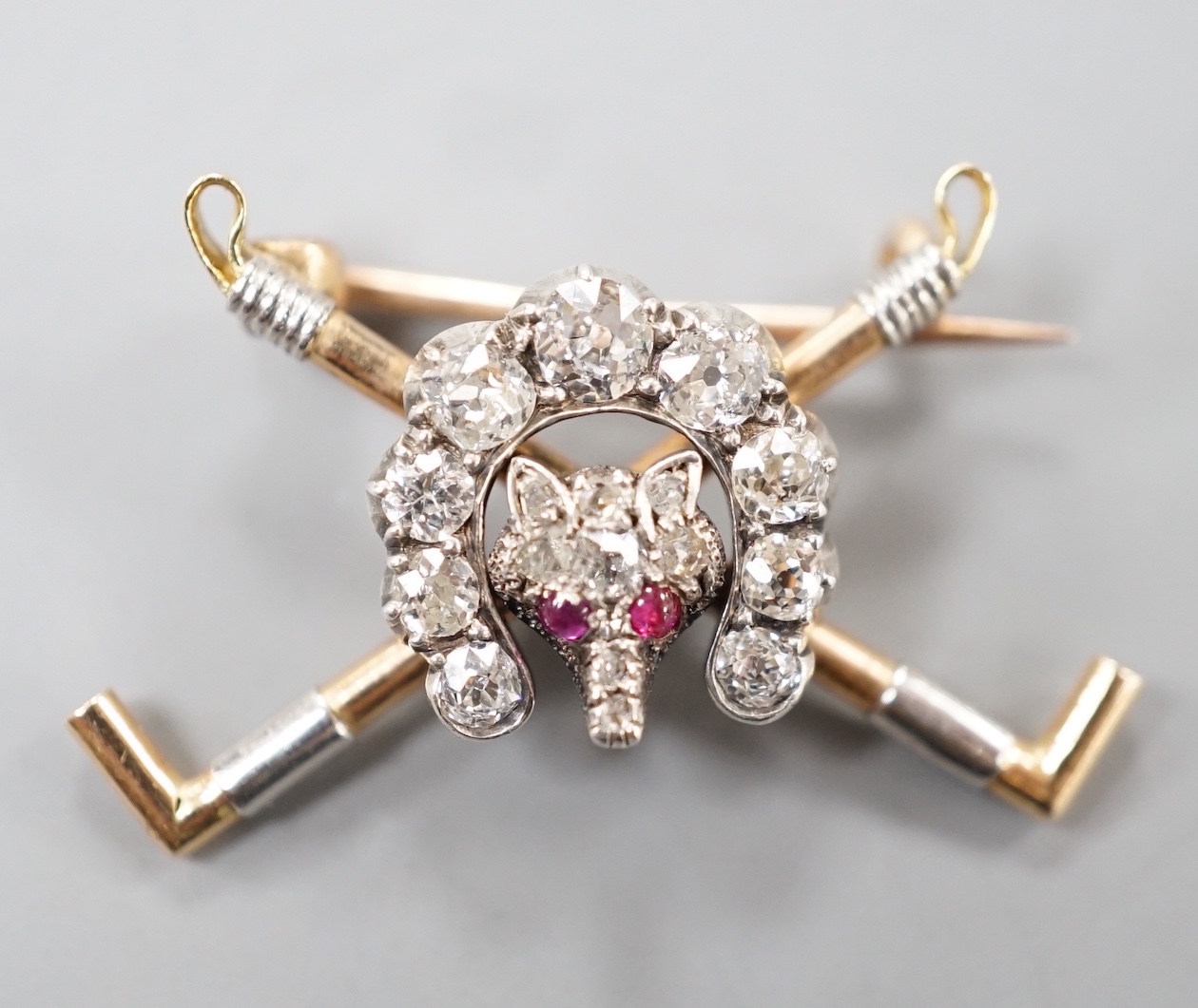 An Edwardian yellow metal, ruby and diamond set hunting brooch, modelled as a horseshoe and fox head upon crossed riding crops, 29mm, gross 4.2 grams.